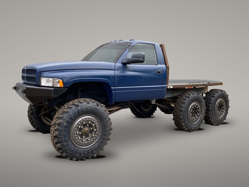 The Dodge Off Road crew's blue 1998.5 Ram 2500 regular cab 6x6 with its steel flatbed and headache rack, on three axles with big beadlock wheels and mud tires, shown against a plain gray studio-style backdrop (background edited)