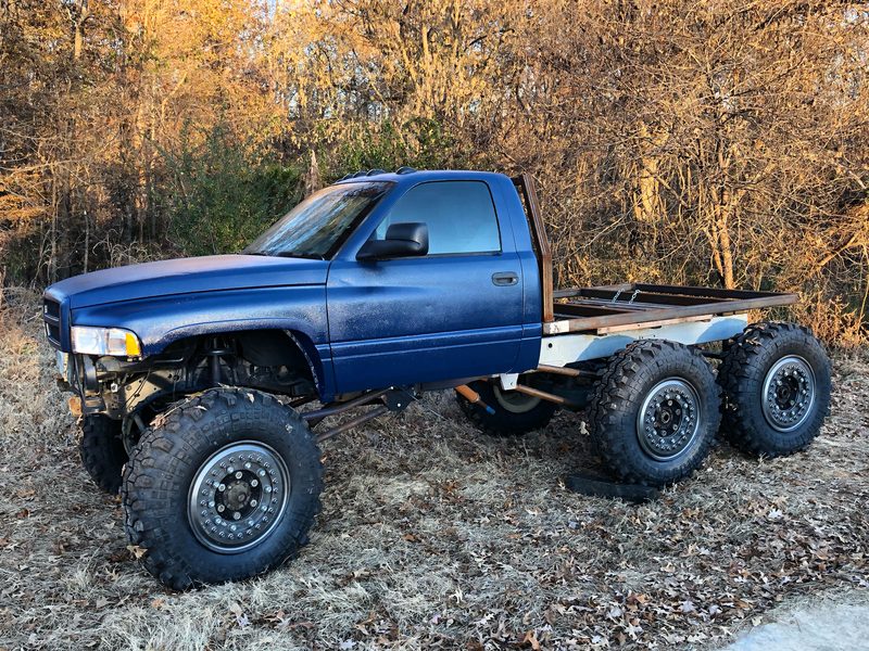 Front three-quarter view of the blue Ram 6x6 in autumn woods, showing the long-arm links and coilovers under the front