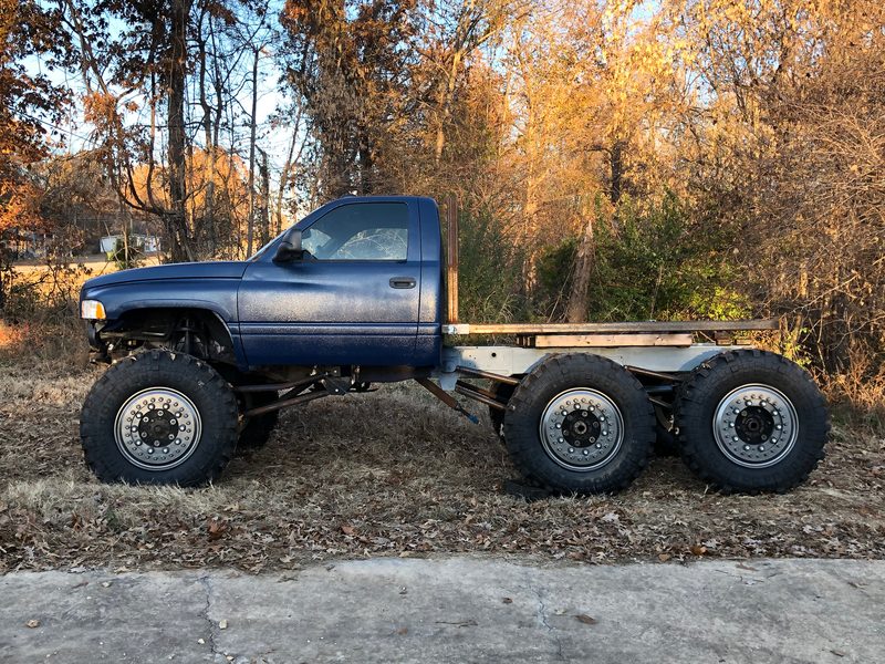 Side profile of the blue Ram 6x6 parked in autumn woods, with a bare bed frame on the back and three axles on big beadlock wheels